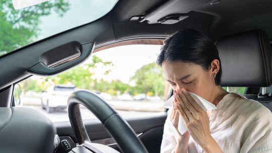 Unwell Asian female driver sneezing into a paper tissue in her car. Health problem, allergy, and sick driving concept.