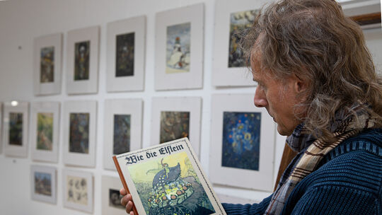 Galerist Wolfgang Diez hält vor den Originalen das Märchenbuch von Auguste Langbein-Mährlen in den Händen. Fotos: Jean-Luc Jacqu